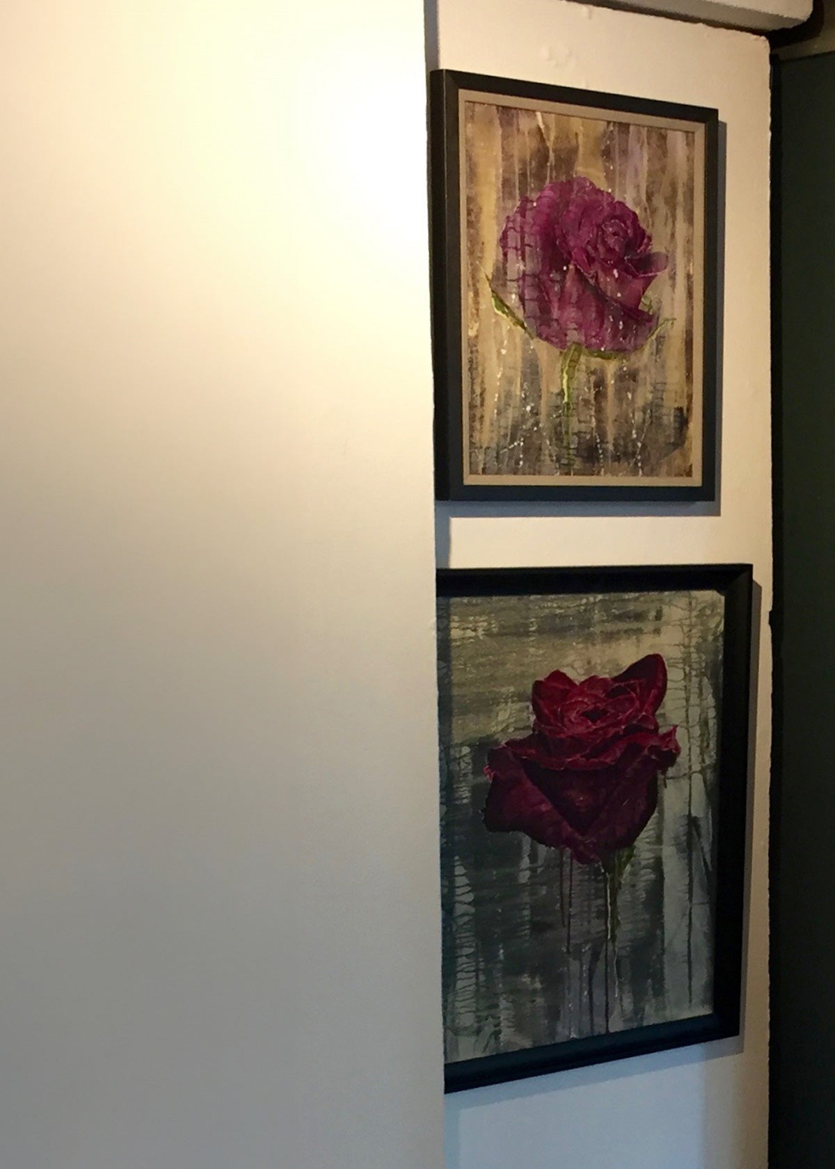 Two framed wall art paintings featuring stylised roses, displayed vertically on a wall. The top print shows a purple rose on a textured beige background, and the bottom print shows a crimson rose on a gray-green background.