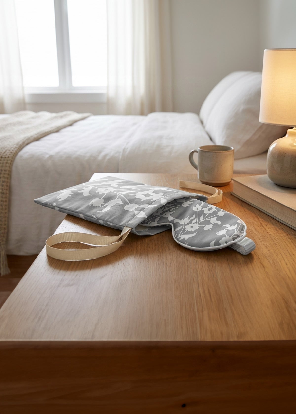 Gray with white floral pattern silk eye mask in its matching bag on a wooden surface, with a bed and lamp in the background.
