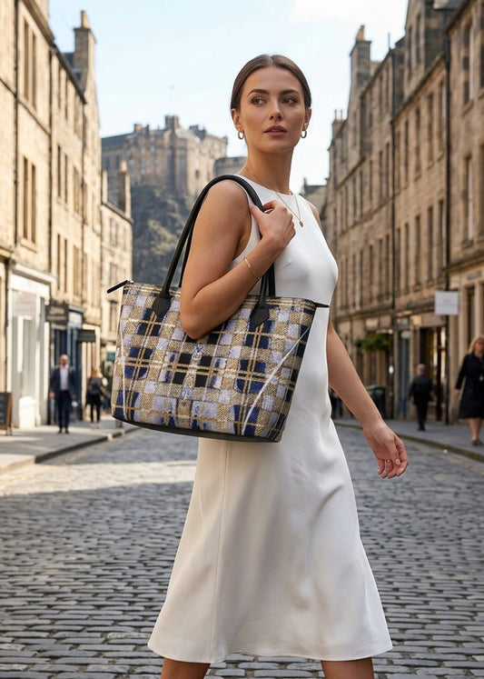 A woman in a white sleeveless dress carries a large woven canvas tote bag with a blue, gold, and beige plaid pattern and black leather handles, walking down a cobblestone street with historic buildings in the background.