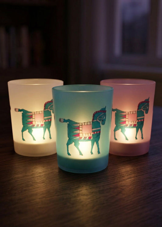 Three frosted glass candle holders with horse silhouettes and patterned fillings, glowing with warm light on a dark wooden table.