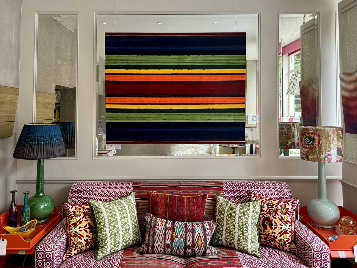 A colourful striped original oil painting titled Stripes Chakra by Rose Long, displayed in a showroom, with a patterned sofa, decorative pillows, side tables, lamps, and mirrors around it.