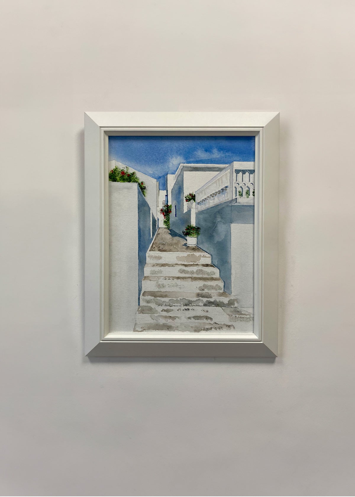 A framed acrylic painting of a Mediterranean-style staircase leading up between white buildings under a blue sky, displayed on a light gray wall.