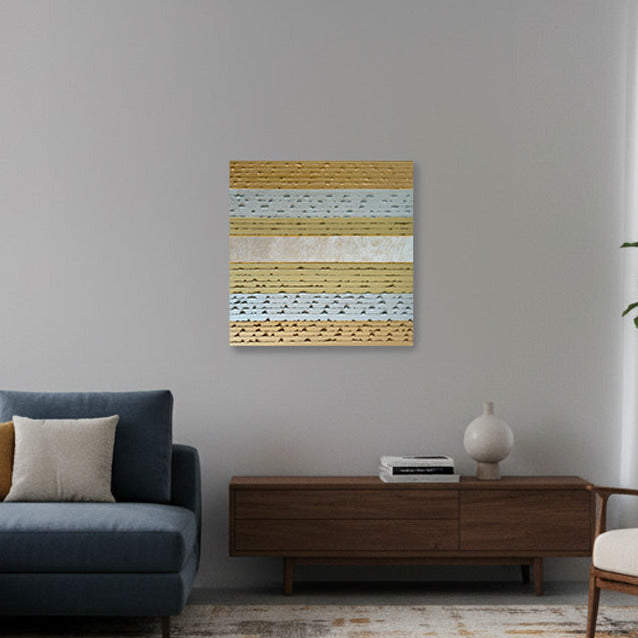 A modern living room featuring a blue sectional sofa with accent pillows, a mid-century style armchair, a wooden sideboard, and a large abstract patterned area rug. A small gold striped square canvas oil painting with gold leaf is mounted on the gray wall above the sideboard.