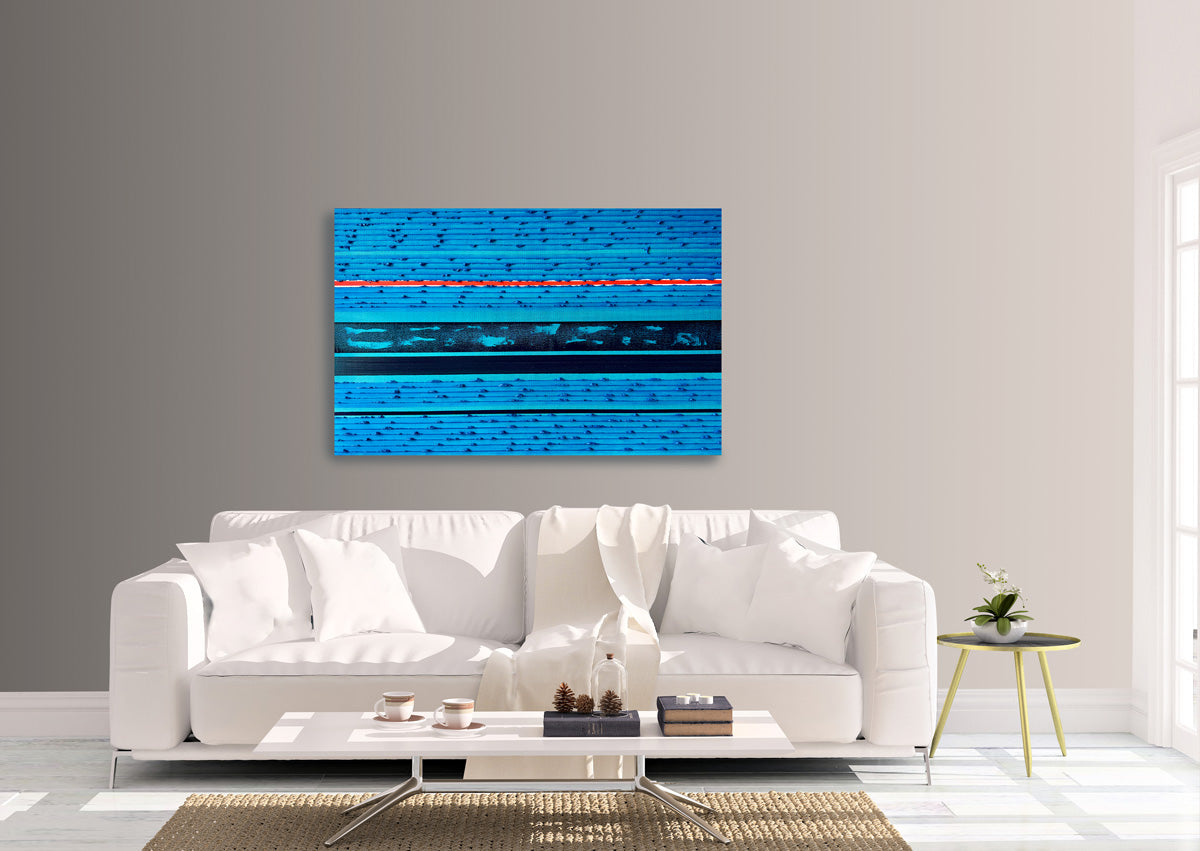 A modern living room setting featuring a square abstract canvas painting with horizontal blue and navy stripes and a thin orange line, hung on a gray wall above a white sofa.