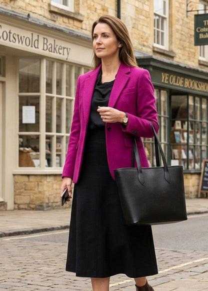A woman stands on a cobblestone street wearing a bright magenta blazer over a black top and black midi skirt, carrying a large black leather tote bag. Shops and stone buildings are visible in the background.