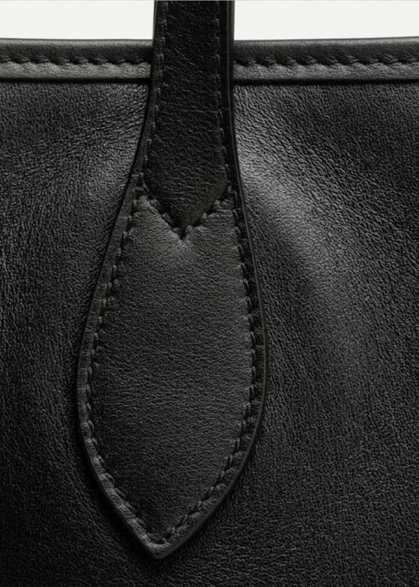 Close-up detail of a black leather bag showing a stitched leather tab and fine grain texture.