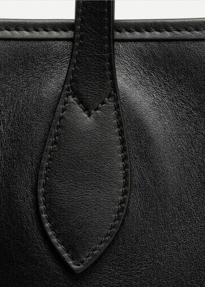 Close-up detail of a black leather bag showing a stitched leather tab and fine grain texture.