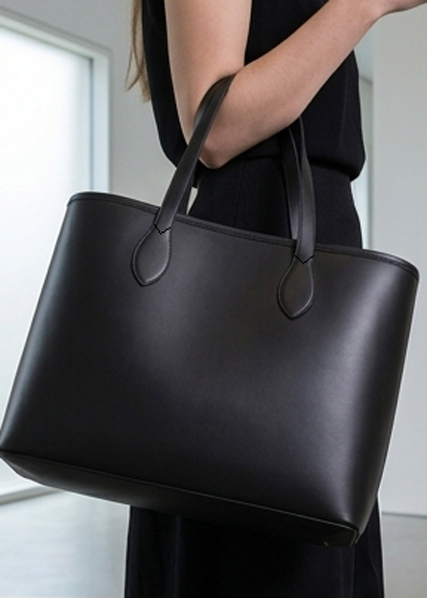 A large black leather tote bag with double shoulder straps, carried by a person wearing a black dress in a bright, modern indoor setting.