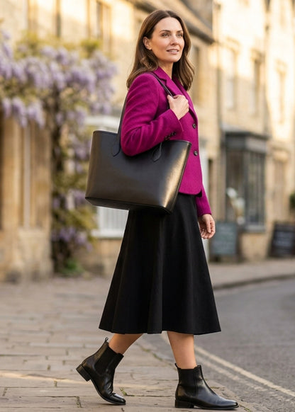 A woman walking on a cobblestone street wearing a bright magenta blazer, a black midi skirt, black ankle boots, and carrying a large black leather tote bag.