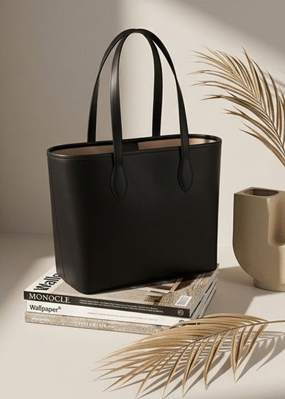 A structured black leather tote bag with double shoulder straps, styled on top of a stack of books next to a beige ceramic vase and dried palm leaves.