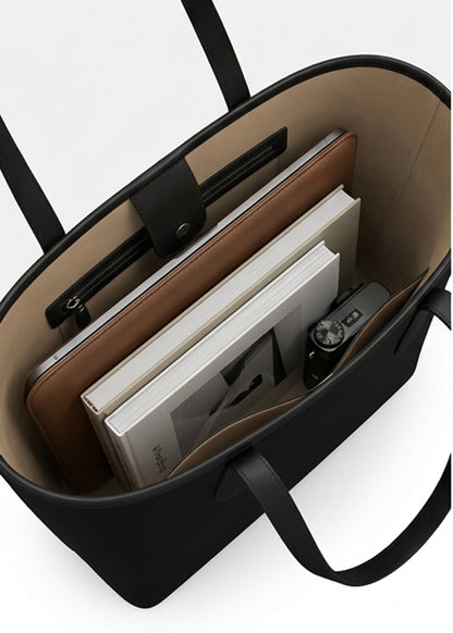 Black tote bag shown from above, filled with books, a brown folio, and a camera, on a white background.