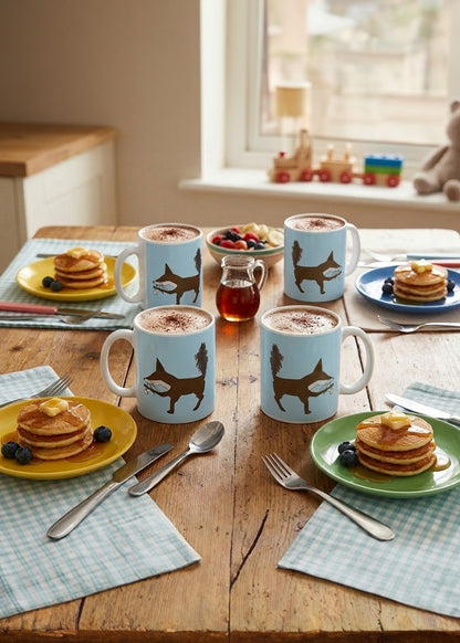 Table setting with blue mugs featuring fox designs, pancakes, and fruit.
