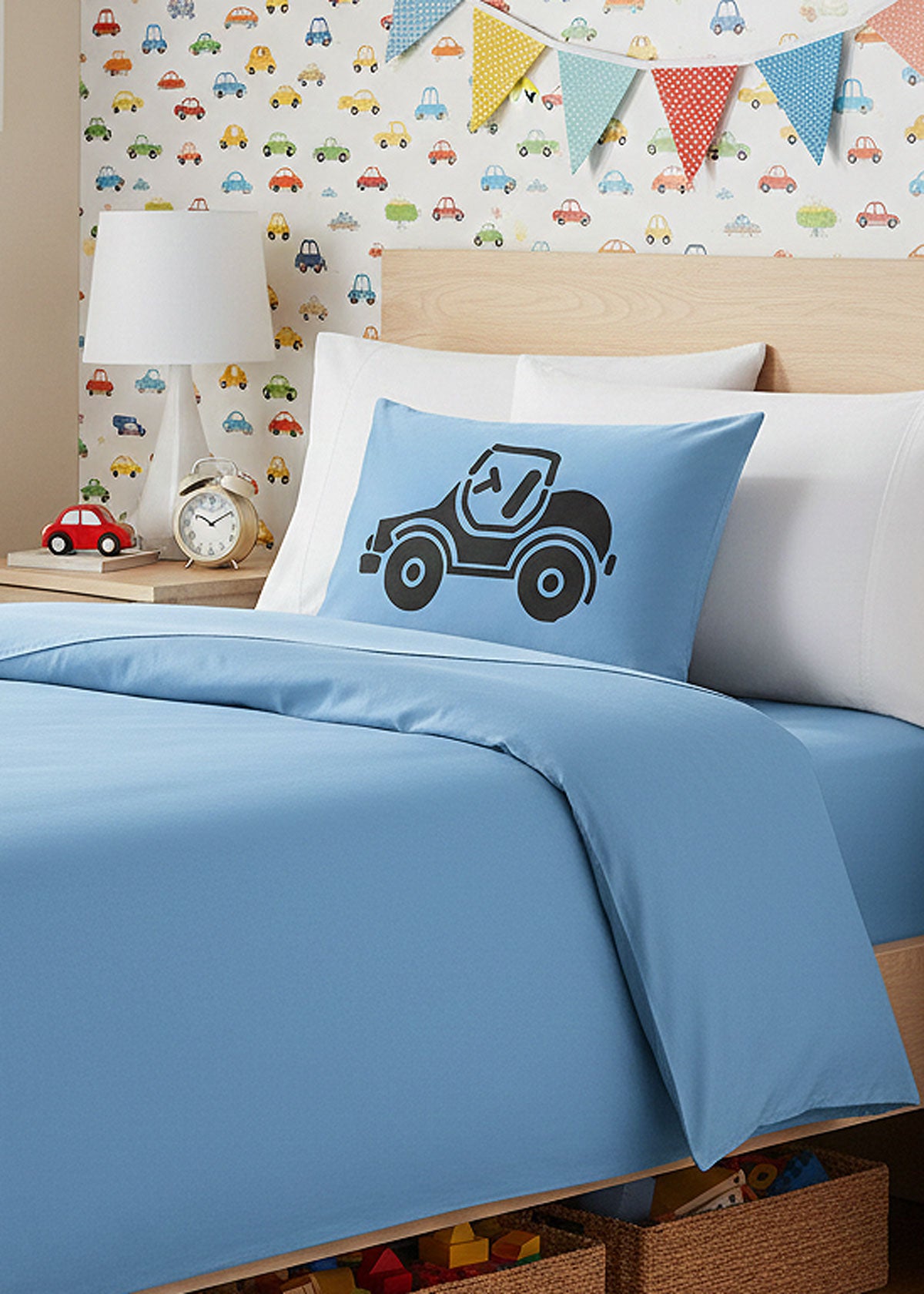 Children's bedroom with blue bedding featuring a blue silk pillowcase with a car design, white pillows, and colorful car-themed wallpaper.