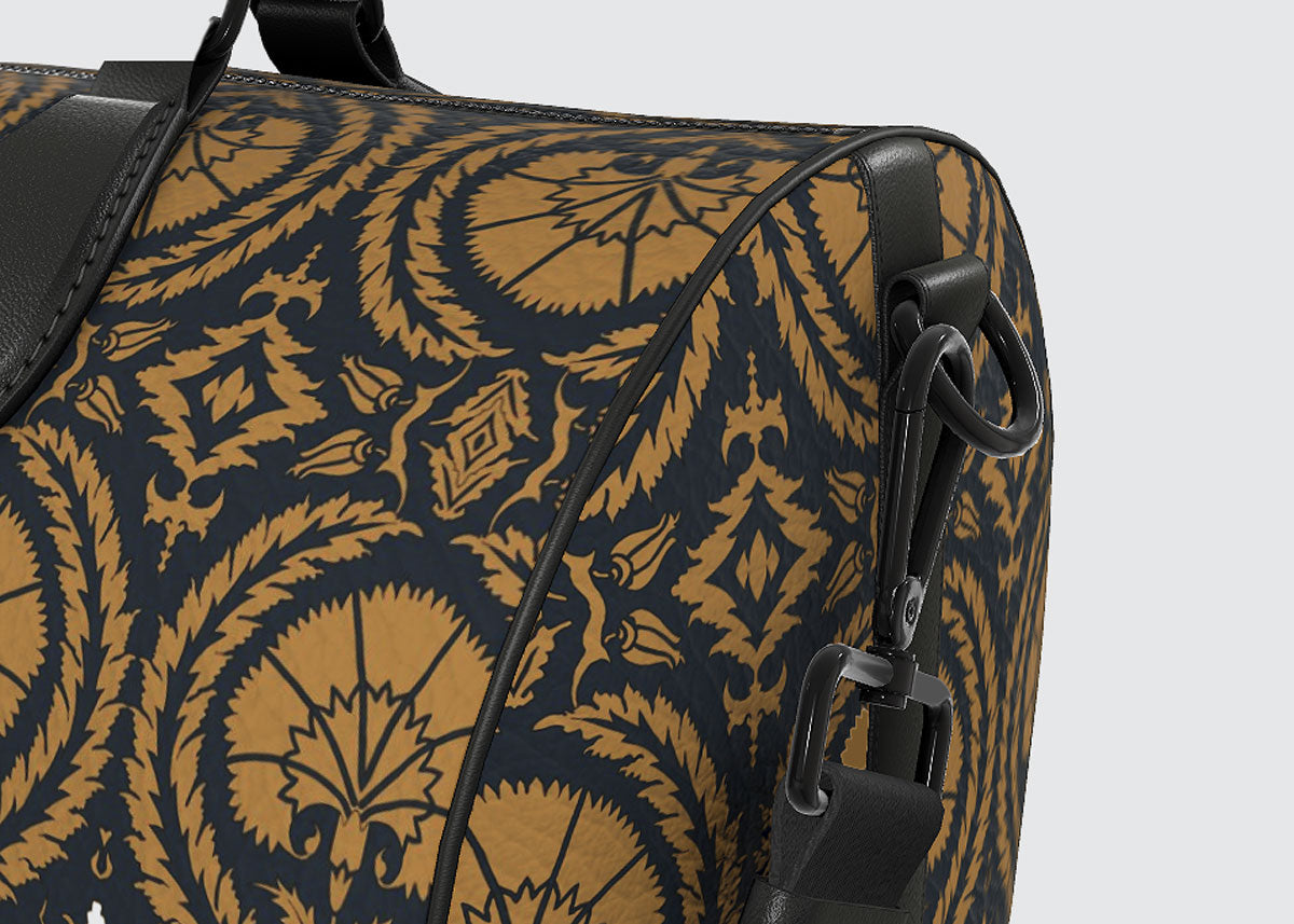 Close-up of a duffel bag featuring a dark navy blue and golden-brown ornate patterned fabric with black leather trim and hardware.