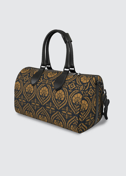 A cylindrical duffle bag with a navy and ochre Suzani pattern, featuring black leather handles and trim, photographed on a light gray background.