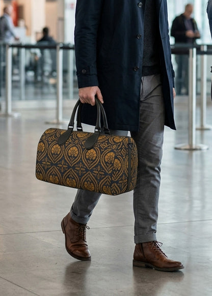 A man in a navy coat, gray trousers, and brown leather boots carries a patterned duffel bag with a dark blue and gold geometric design in a modern indoor setting.