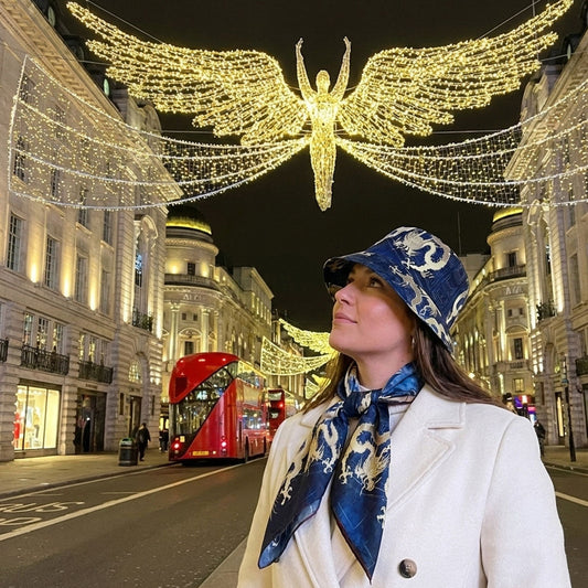 Woman in a white coat and blue patterned bucket hat with dragon motifs, standing under a large decorative light installation in a city street at night.