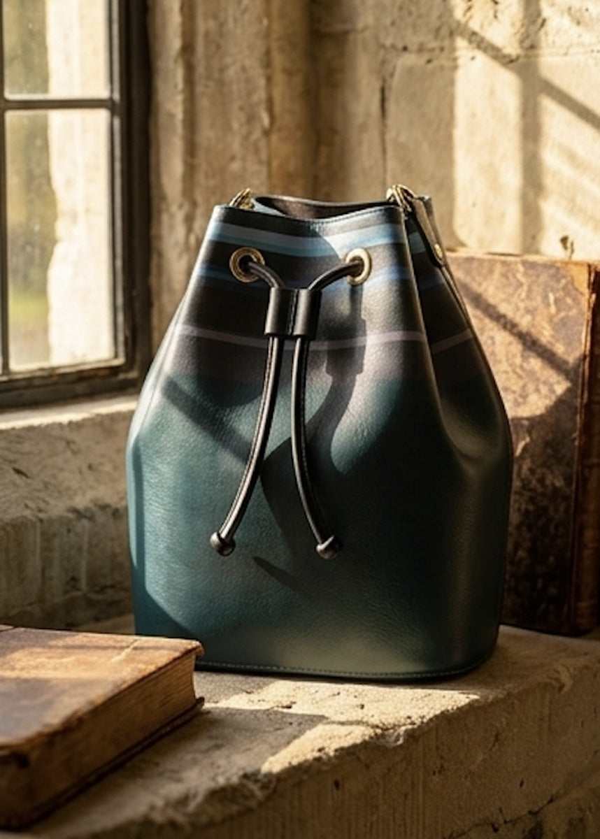 A dark teal leather bucket bag with a drawstring closure sits on a stone windowsill in warm, natural sunlight.