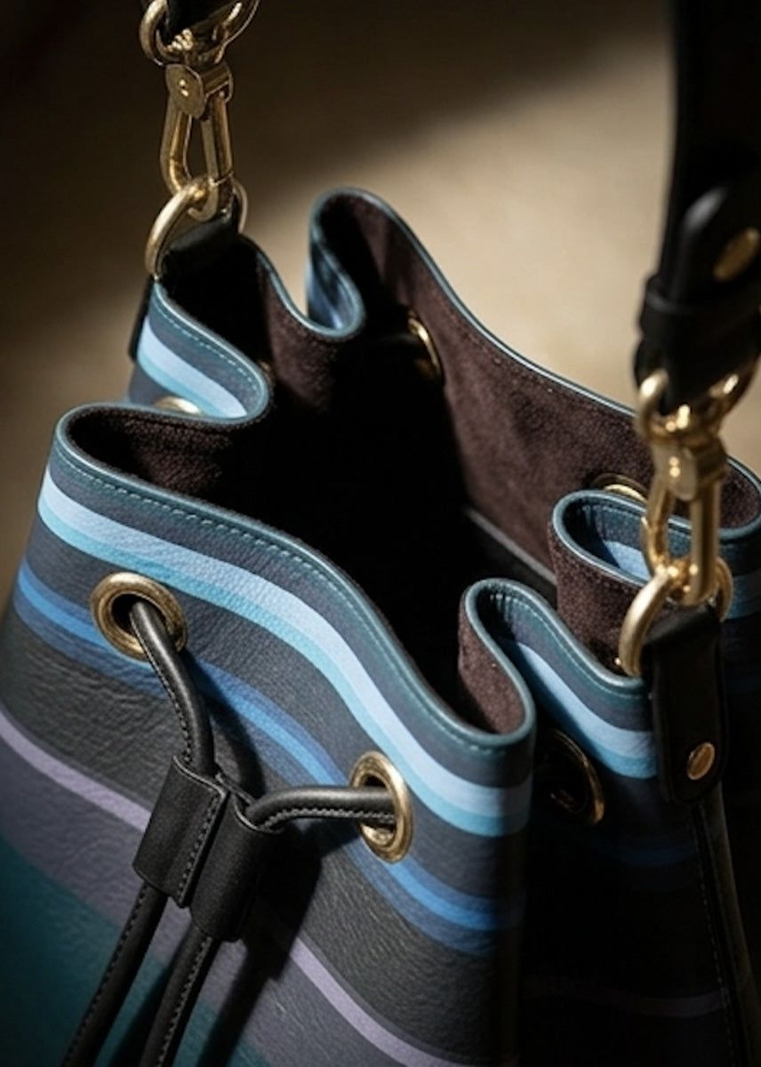 Close-up of a bucket-style handbag with a striped pattern in shades of blue, gray, and black, featuring gold-tone hardware and a faux suede interior lining.