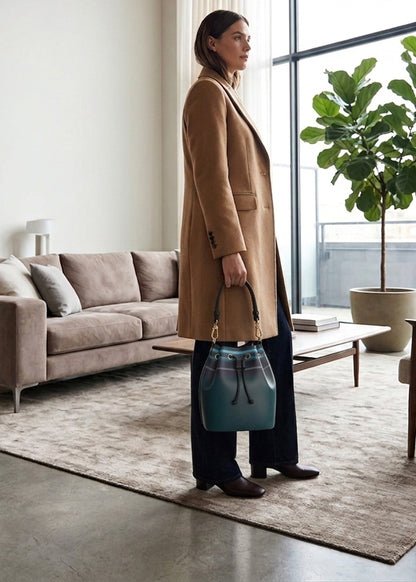 A woman stands in a modern living room holding a teal blue and gray drawstring bucket bag. She is wearing a long tan coat and dark trousers.