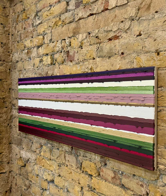 A rectangular original art piece with horizontal multicolor painted textured stripes, mounted on a rustic yellow brick wall.
