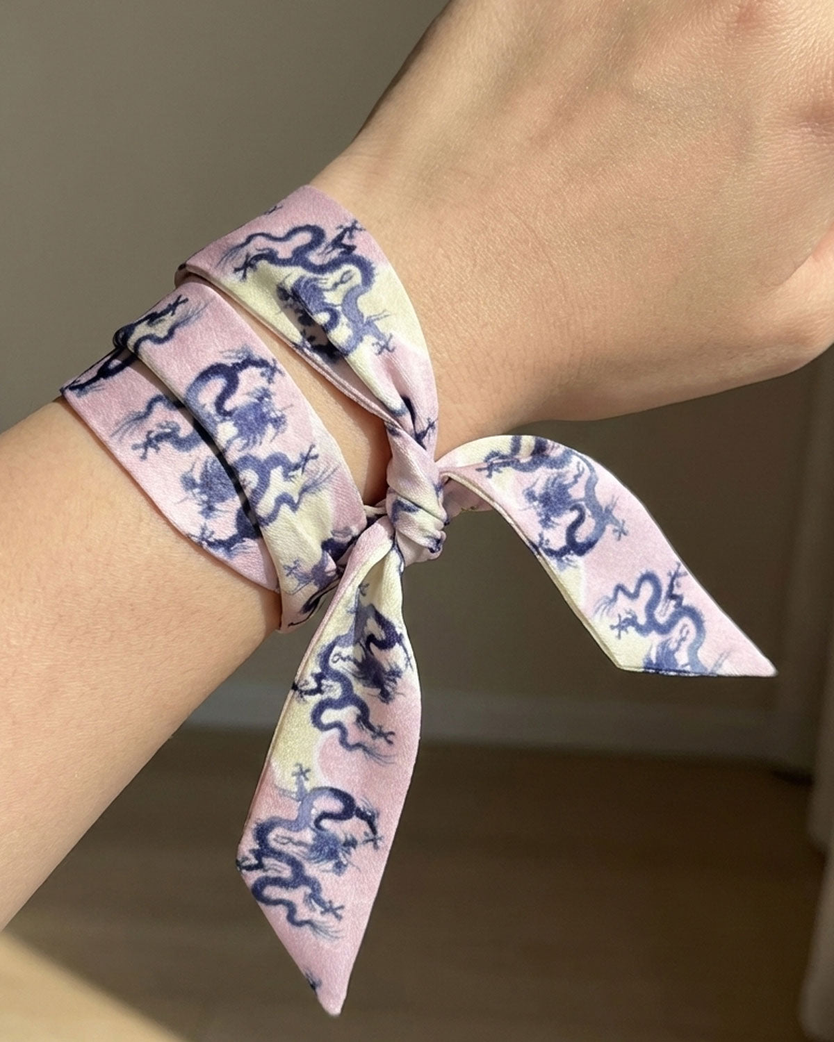 Person with a skinny silk scarf tied around a wrist with blue dragons on a pink and cream background.