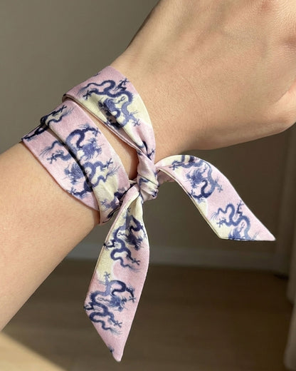 Person with a skinny silk scarf tied around a wrist with blue dragons on a pink and cream background.