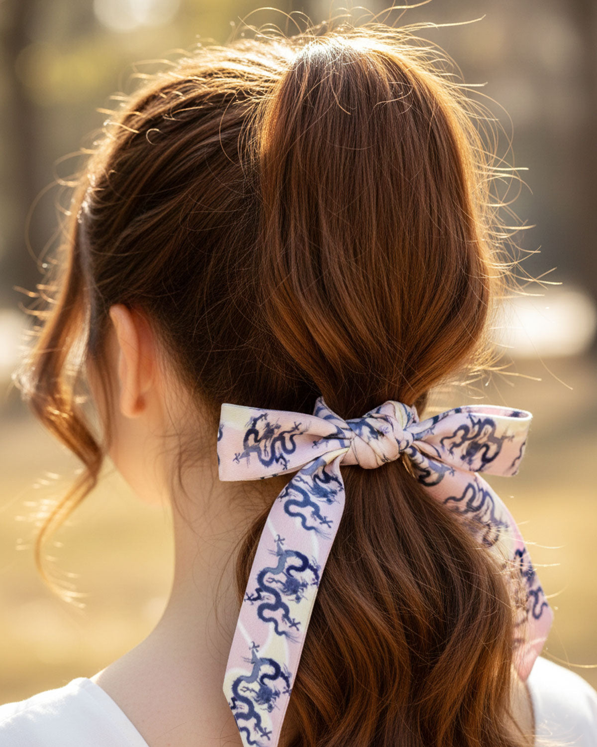 Person with a ponytail tied with a decorative Skinny hair silk scarf with blue dragons on a pink and cream background.