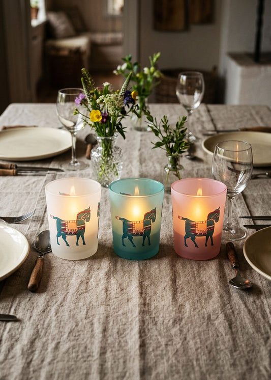 Three lit candle holders with a geometric horse design in white, teal, and pink, arranged on a linen tablecloth with dinnerware and flowers in the background.