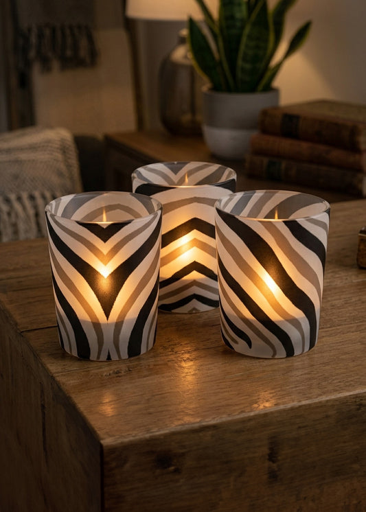Three lit candle holders with a black, white, and gray zebra pattern, arranged on a rustic wooden table in a cozy, warmly lit room.