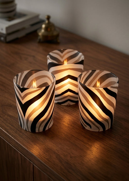 Three zebra-print glass candle holders with lit candles, arranged on a dark wooden table in a warm indoor setting.