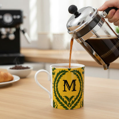 Person pouring coffee from a French press into a mug with a yellow and green wreath design and letter 'M'.