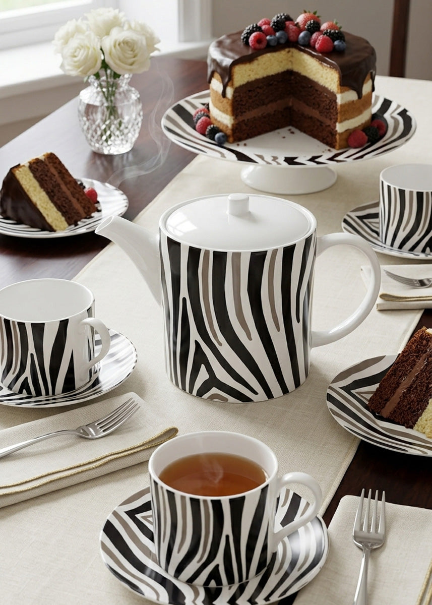 A zebra-print ceramic teapot and matching cups and saucers arranged on a table, alongside a chocolate cake on a stand and a vase of white flowers in the background.